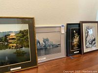 Four framed pieces of wall art standing on floor against wall: temple photo with bamboo frame, iceberg photo in gray matte, silk embroidered floral in black frame, panda artwork in brown frame.