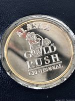 Close-up of gold colored medallion embossed with 'ALASKA GOLD RUSH CENTENNIAL' and prospector image