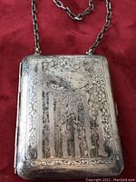 Front view of antique rectangular silver plated purse with chain on red background, showing detailed engraved patterns and central oval cartouche.