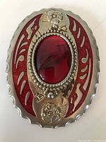 Front view of oval belt buckle showing large red glass center and surrounding red enamel with metal floral details.