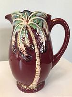 Front view of burgundy ceramic pitcher with raised palm tree design in gold and green.
