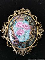 Clear, full view of the brooch showing the oval porcelain medallion with pink cabbage roses and detailed gold scroll pattern on background, framed by textured gold-tone metal ribbon edging.