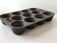 Angled view of the cast iron muffin pan showing 11 deep holes and flat rim with holes for hanging, surface shows patina and wear.