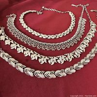 Four silver tone Coro necklaces laid out on red fabric, showing varied detailed patterns and lengths.