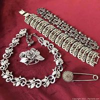 Full lot on red fabric showing Coro brooch with roses, two silver tone bracelets, leaf vine necklace and large decorative safety pin kilt pin.
