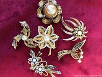 All six vintage damascene brooches displayed on dark red fabric. Each brooch has gold and black damascene detailing with faux pearls in various floral and leaf designs.
