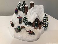 Front and side angle showing the detailed snow-covered house, brown brick chimneys, decorated Christmas tree with multi-colored lights, and four small children sledding on a glittery snow base.