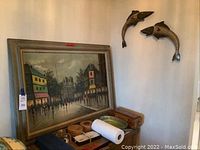 Wide view showing framed cityscape painting resting on furniture below two metal fish sculptures on the adjacent wall, along with small decorative items on the furniture surface.