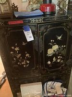 Front view of black Asian-style cabinet with gold floral and bird artwork on doors and ornate metal door handles.