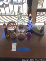 Full view of the eight glass and ceramic decorative items on a table under sunlight. Included are a large clear glass elephant missing an ear, two ceramic half-doll pincushions, several glass paperweights, and glass animal figurine.