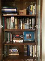 Books displayed in a glass cabinet with multiple shelves showing spines of books including both hardcover and paperback types, arranged vertically and horizontally.