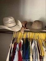 Wide shot of multiple cowboy hats on shelf and assorted clothes on rack beneath with yellow hangers.