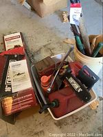 Photo of Craftsman 16-inch electric chainsaw in white container, gas can, bolt cutters, manual, and hedge trimmers in bucket on concrete floor, showing all items in lot.