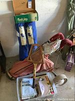 Photo showing pinkish golf bag with golf clubs, vintage wooden Wilson tennis racket leaning on it, golf towels and balls in plastic container, with blue folding chairs in background.