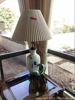 Full view of the lamp with ceramic crane base and beige pleated shade on glass table near window