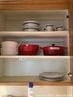 Kitchen cabinet shelf showing Le Creuset red baking dish, red mixing bowls, white ribbed bowls, and cups with saucers