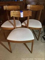 Three mid century wooden chairs with white vinyl seats arranged side by side. Shows the full shape and style of the chairs with wood backs and seat frames.
