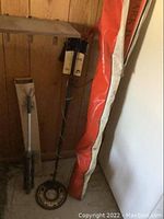 Side view of skis in their bag and the metal detector upright beside the wooden tripod against a wall.