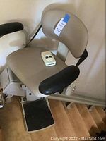 Front view of beige padded stair lift chair mounted on rail with footrest and remote control on seat.