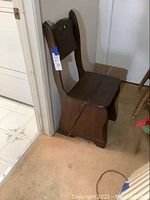 Side view showing the chair folded in sitting position against wall and door frame, dark wood finish, visible wear on carpet beneath