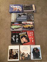 Two sealed 1000-piece animal jigsaw puzzles and 9 assorted books arranged on a beige carpet. Books cover topics including Bateman birds, Ford cars, Jeep trucks, Beatles, Elvis Presley, and Norman Rockwell art. The puzzle boxes are sealed and in good condition.