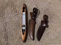 Photo showing wooden knife sharpener with attached thermometer and two knives with leather sheaths.