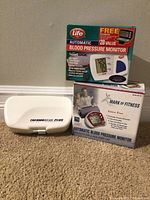 Set showing two blood pressure monitors in their boxes and a white Thermoscan Plus thermometer case on carpeted floor.