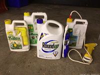 Four containers of weed and brush control products including Killex, Roundup, and Pathclear arranged on the ground showing sizes, labels, and spray attachments.