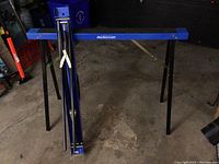 Two metal sawhorses with blue crossbars and black foldable legs, standing on concrete floor showing used condition.