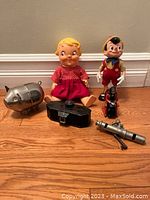 Group shot of vintage Pinocchio doll, vintage sitting doll in red, metal pig sculpture, black binoculars, scooter figurine, and metal cannon figurine on a hardwood floor against a beige wall.
