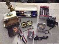 Various used miscellaneous items laid out on a table including binoculars, watches, cutlery set, brass scale, and small boxes.