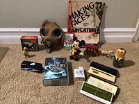 Wide view of all lot items arranged on floor showing Lego box, caricature book, Cross pen and pencil boxes, figurines, Twilight Zone Blu-ray, and gas mask.