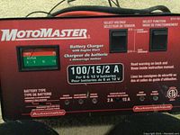 Front view of MotoMaster battery charger showing voltage selector switch set to 6V, function mode set to charge. Analog ammeter visible on left side. Model and branding printed prominently.