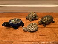 Four turtle figurines displayed on a wooden floor, showing different styles and materials for each turtle.