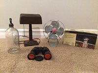 Photo showing entire lot including the glass seltzer bottle with metal siphon head, brown desk lamp, small metal vintage fan with red center label, binoculars, and unopened box with outdoor lamp image.
