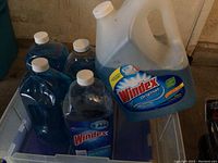 Five bottles of Windex Original glass cleaner in a plastic tub, four smaller spray bottles and one large gallon jug showing front label.