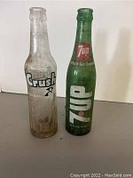 Both vintage glass soda bottles shown upright on a flat surface: a clear Crush bottle and a green 7-Up bottle, both with original logos and visible dirt.