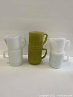 Photo showing 2 avocado green barrel style stackable Fire King mugs with D handles and 4 white Fire King mugs, all stacked in pairs.