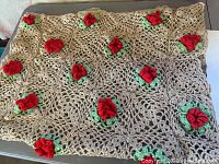 Overall view of the beige crochet throw blanket featuring red flowers with green leaves scattered across the surface