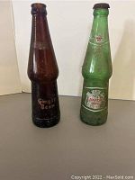 Pair of vintage glass soda bottles, one brown Ginger Beer and one green Canada Dry, both needing cleaning and showing dirt and age.