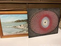 Two art pieces displayed side by side: a circular string art in red, white and blue-grey threads on dark board, and a framed beach scene painting showing waves and trees.