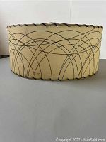 Side view of round beige fiberglass lampshade with black overlapping curved line patterns and black trim stitch detail on edges.