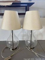 Two matching bedside table lamps with clear glass bases and white textured shades, showing front view including cords.