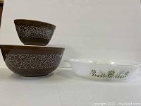 Two brown Pyrex nesting bowls with white floral designs stacked, alongside white Glasbake divided dish with green floral motif.