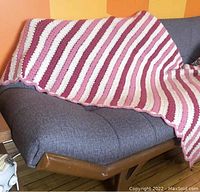 Afghan draped on a grey couch showing full length and striped pattern of pink, white, and burgundy colors.