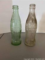 Two vintage Coca-Cola bottles side by side showing clear and green tint with embossed logos.