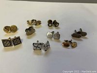 Photo showing nine pairs of vintage cufflinks arrayed in two rows on a white sheet. Various metal finishes, shapes, and decorative patterns visible.
