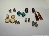 Seven pairs of vintage earrings and one pair of cufflinks arranged on white background showing variety of styles and colors.