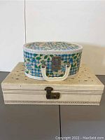 Photo of two jewelry boxes stacked. The top is an oval blue and green floral print box with white handle and metal latch. The bottom is a rectangular cream box with star perforations and latch, both showing stains and wear.