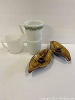Photo showing two leaf-shaped brown glazed Blue Mountain Pottery candle holders and three vintage white glass mugs, one decorated with green floral pattern and two plain.